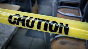 Color photograph of a yellow piece of caution tape stretched out blocking access to an area in a building. The word "CAUTION" is printed in large black block letters on the yellow tape.